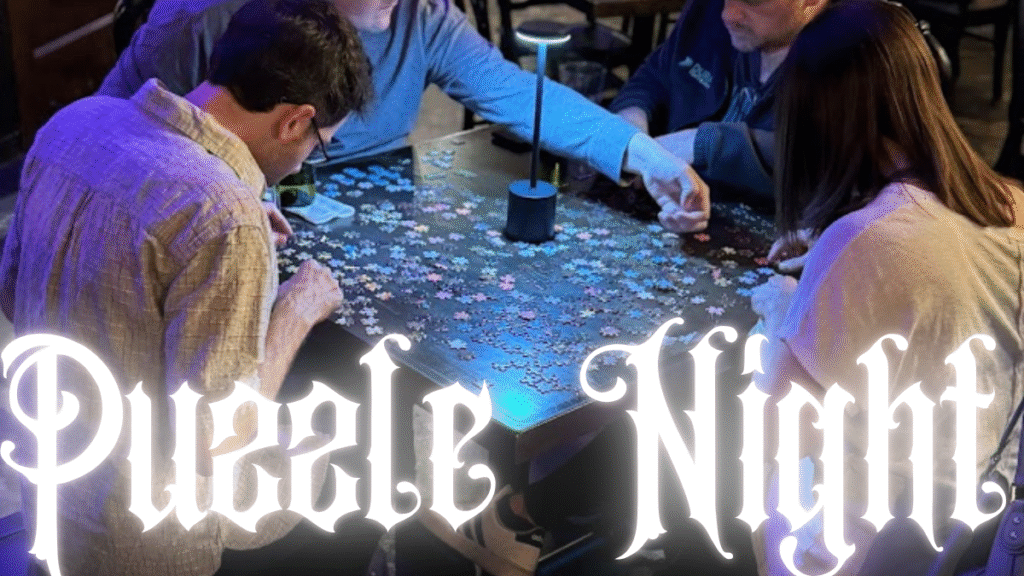 A team of four adults working together to complete a 500-piece puzzle at a lively puzzle night event, with glasses of wine on the table.